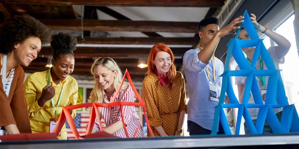 Grupo diversificado de pessoas trabalhando juntas para construir estruturas com cartões coloridos em um ambiente descontraído.