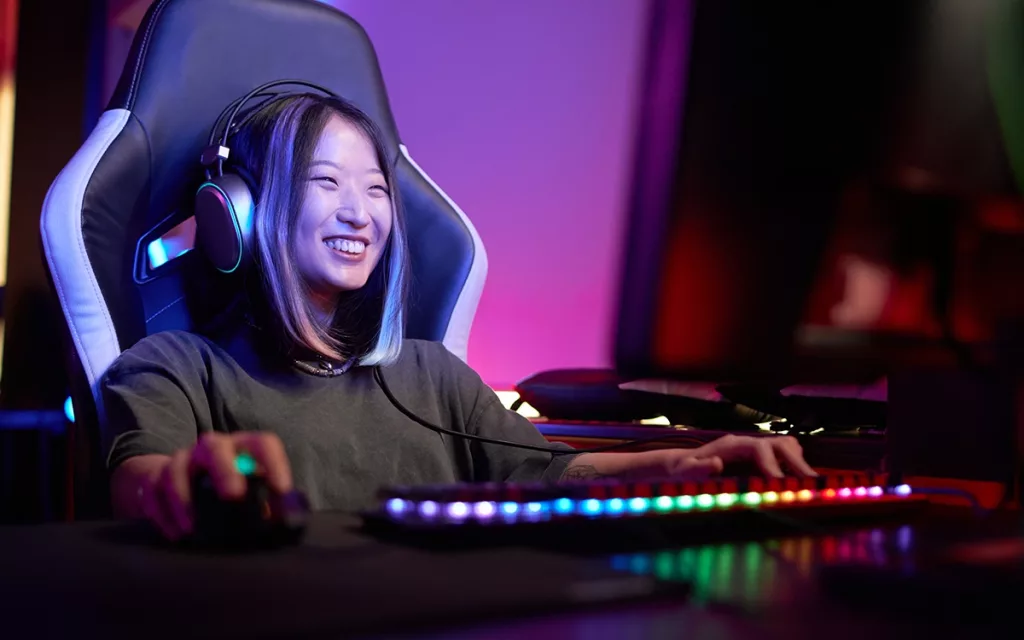 garota gamer sentada sorrindo em cadeira gamer segurando mouse com a mão no teclado jogando game no computador